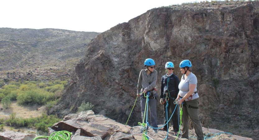 three people in rock climbing gear hold ropes as they stand on a cliff